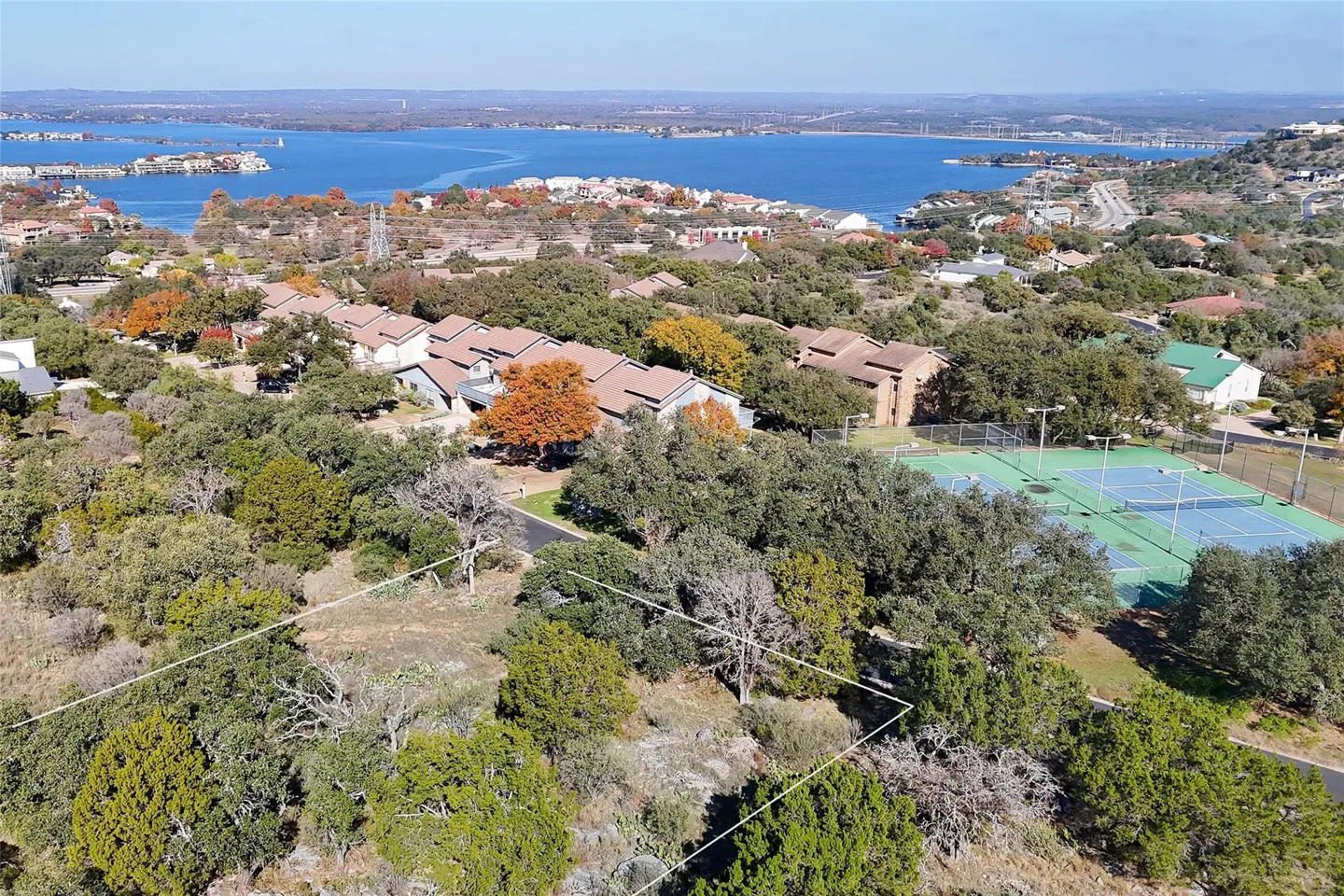 Aerial view of a wooded lot outlined in white, with a lake, houses, and tennis courts in the background.