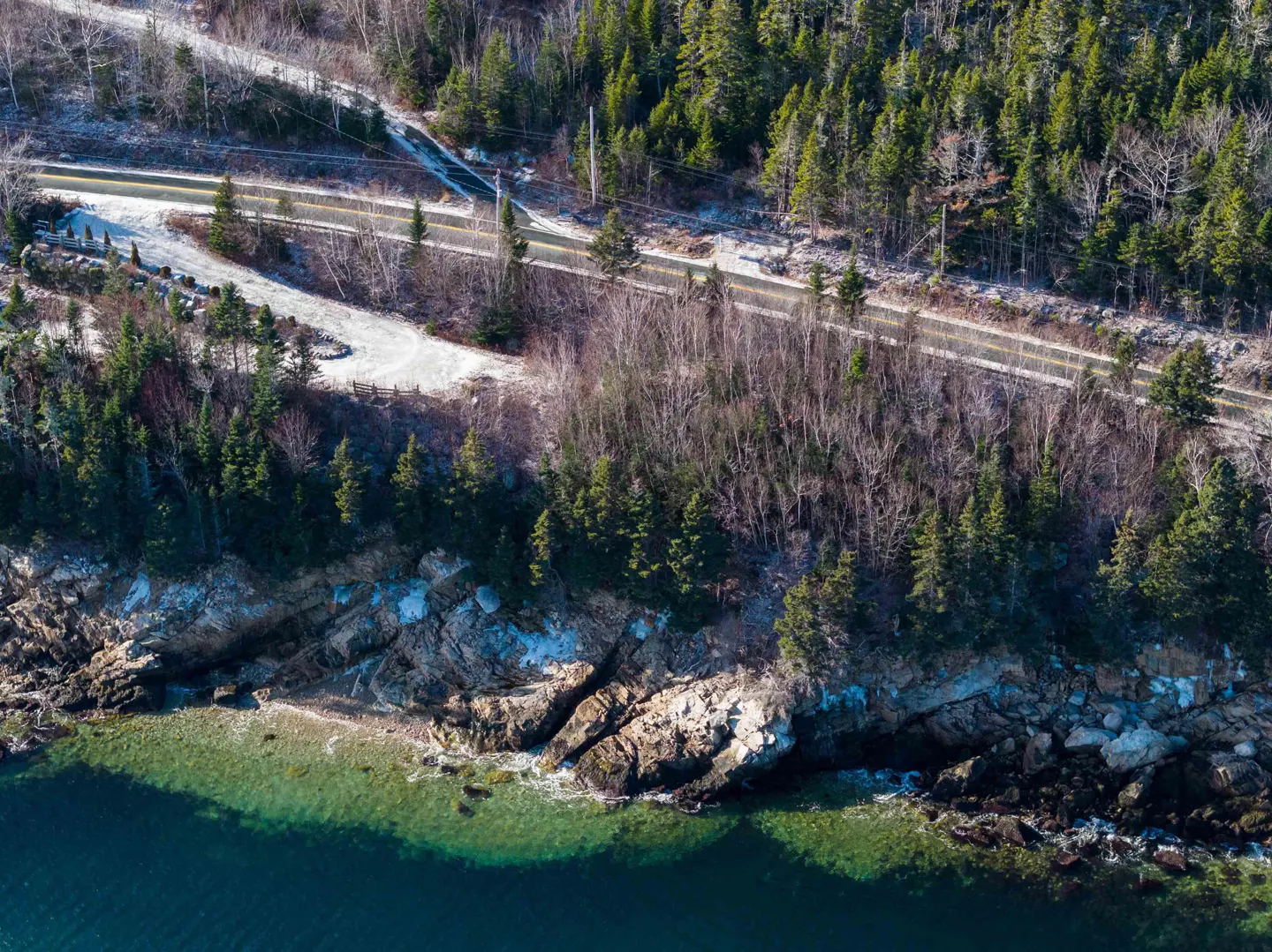 Aerial view of a coastal road bordering a rocky shoreline with evergreen trees and clear blue-green water.