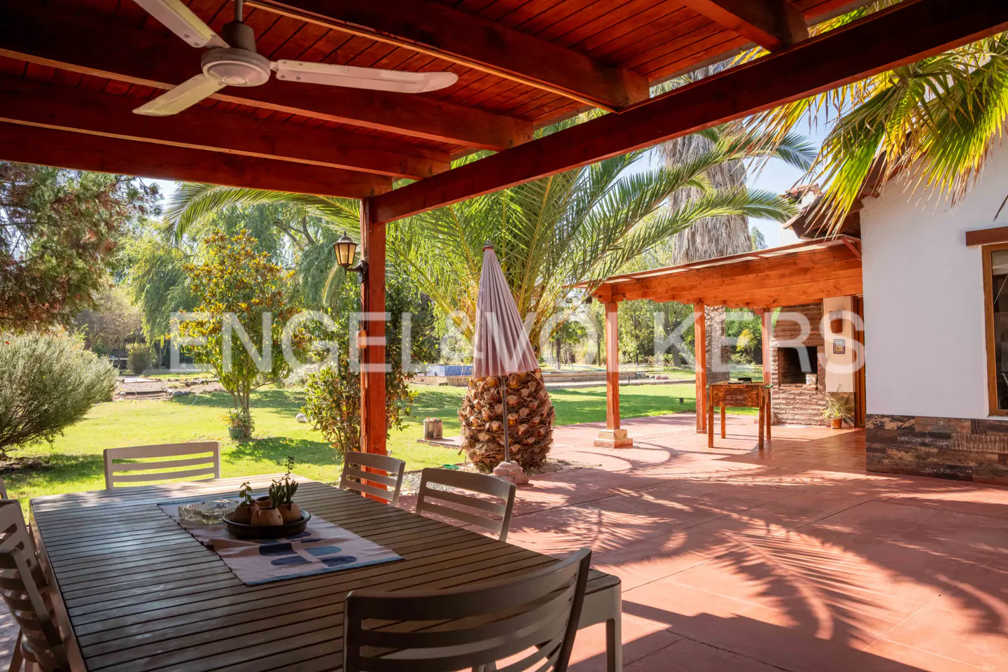 Outdoor patio with a wood table, chairs, and a ceiling fan. A large yard with trees and a building are in the background.