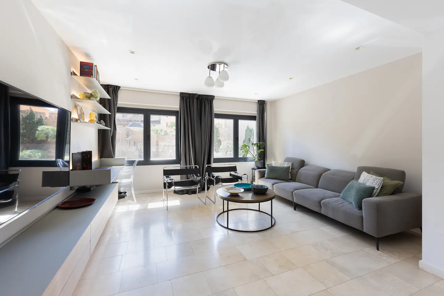 Bright living room with gray sofa, black chairs, and round coffee table. Windows with dark curtains let in natural light. Shelves and a TV are on the left.