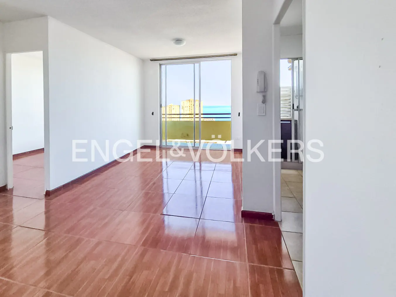 A bright, empty room with white walls and brown tile floors. A sliding glass door leads to a balcony with an ocean view.
