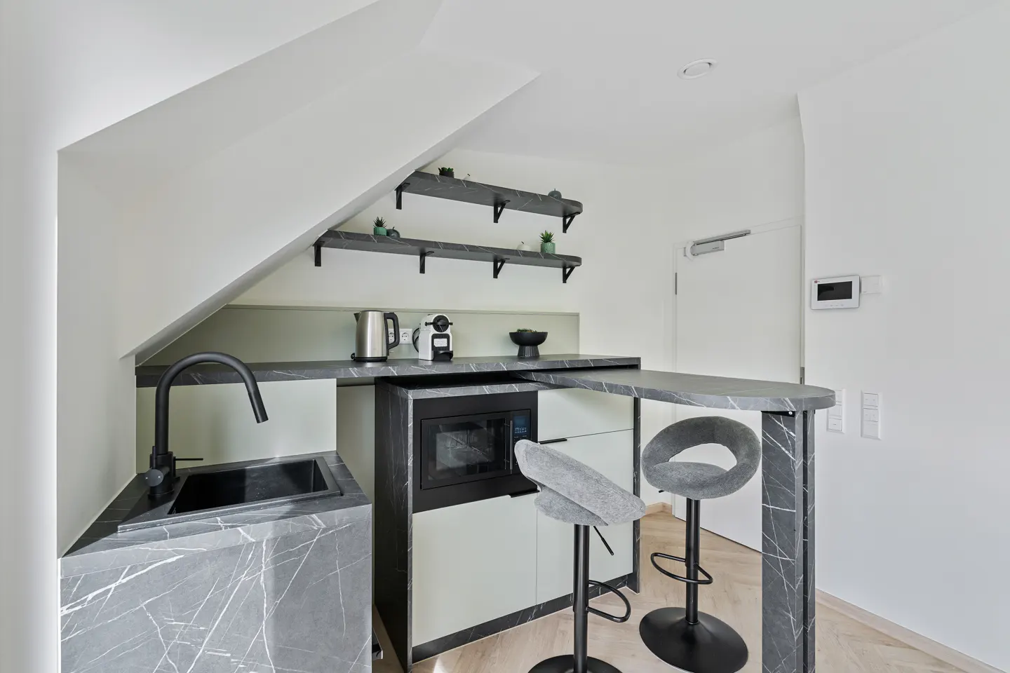 Modern kitchen with gray marble countertops, black sink and faucet, two gray bar stools, and two shelves with plants.