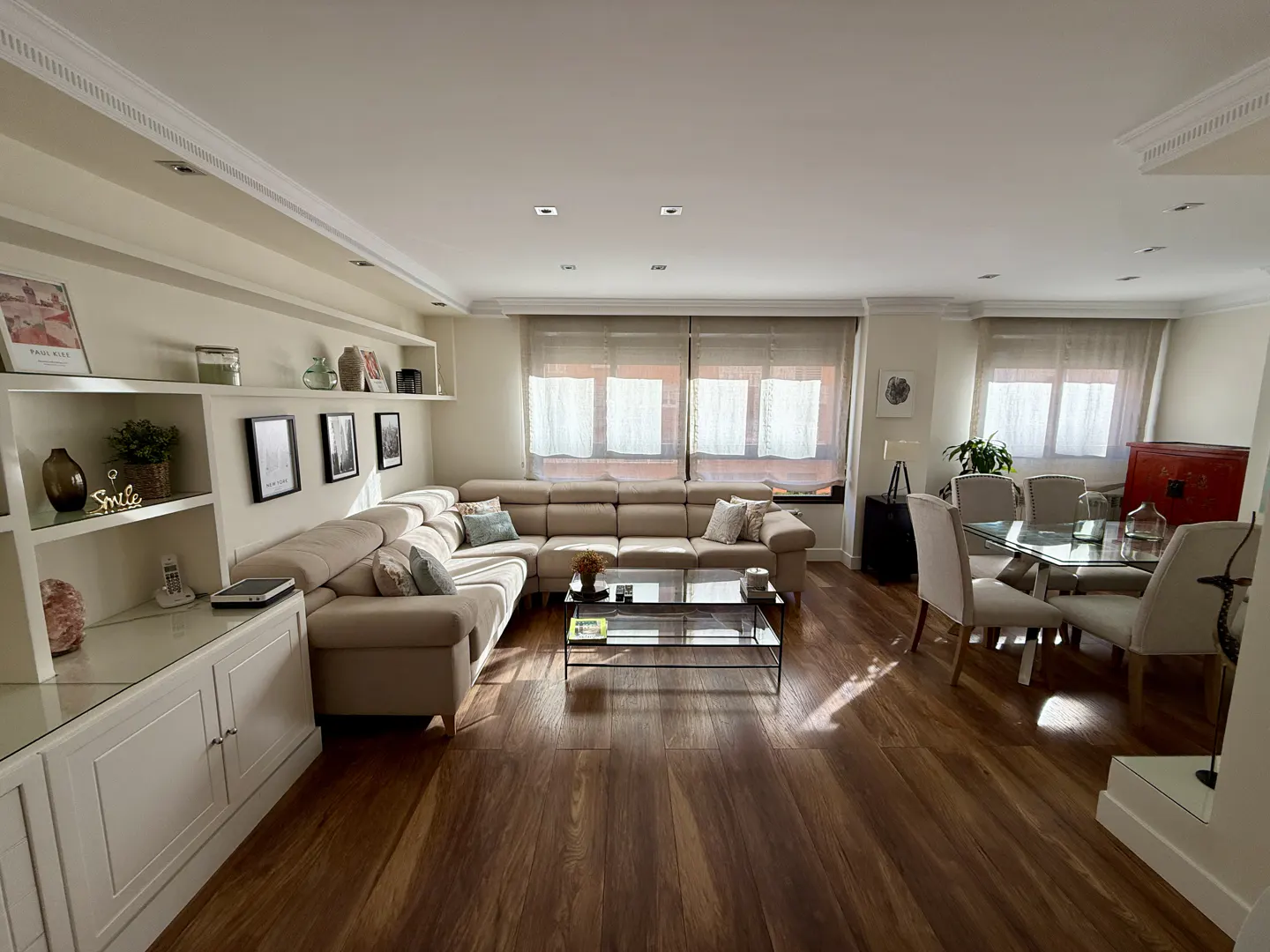Bright living and dining area with hardwood floors. Beige sectional sofa, glass coffee table, and dining set with glass table. Shelving unit on the left.