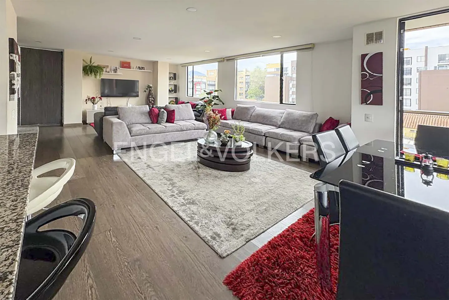 A bright living room with gray sofas, a dark wood floor, and a gray rug. A black dining table is on the right.