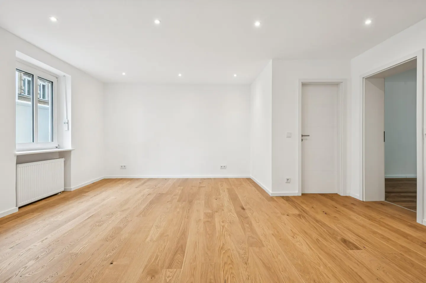 Bright, empty room with light wood floors, white walls, and recessed lighting. A window and two doorways are visible.