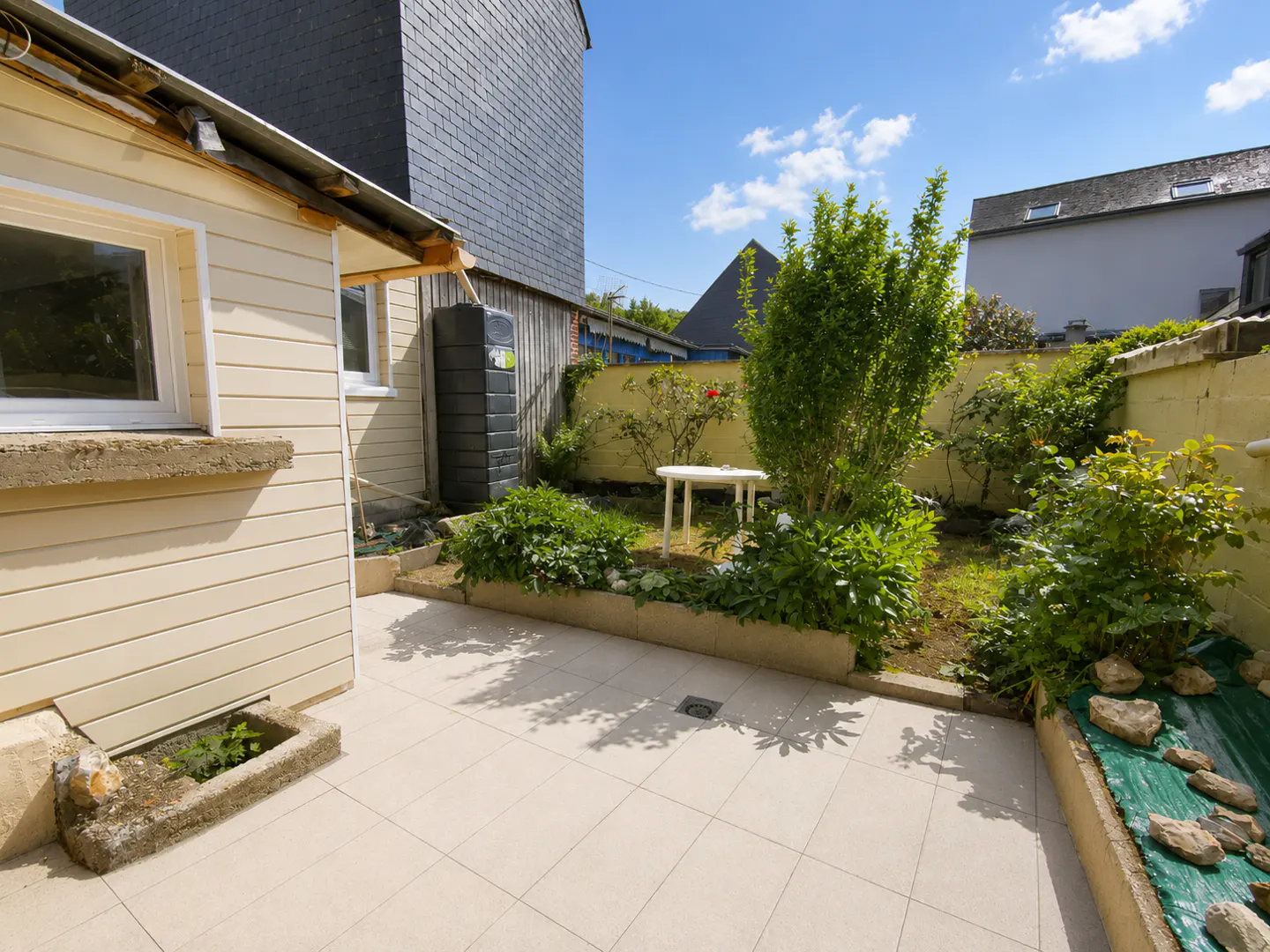 Small backyard patio with light-colored tiles, a white table, lush green plants, and a cream-colored house exterior.