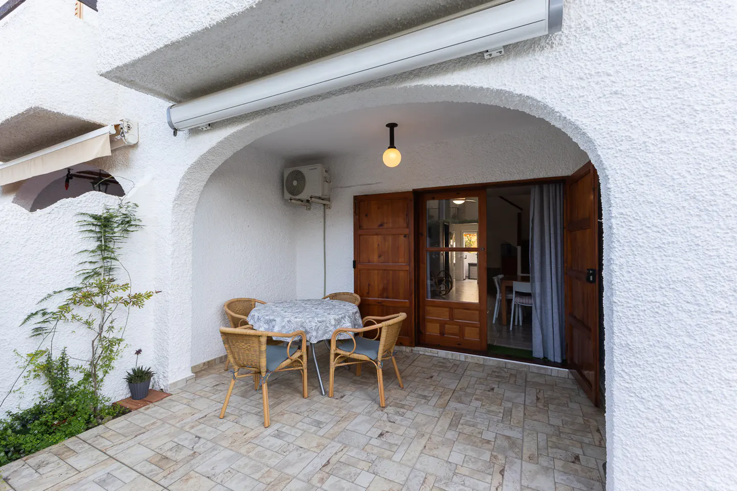Outdoor patio with a round table, four chairs, and open wooden doors leading inside. White stucco walls and tile flooring.