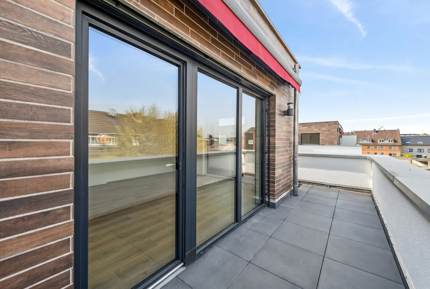 A modern balcony with gray tiles, a white railing, and a brick wall with large sliding glass doors. A red awning is visible above the doors.