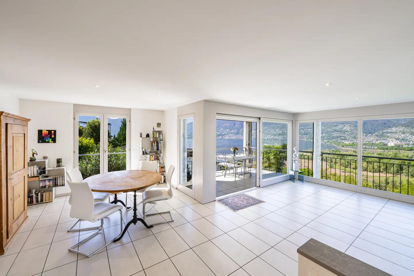 Bright, airy living room with white tile floors, a wood table, and a balcony overlooking a lake and mountains.