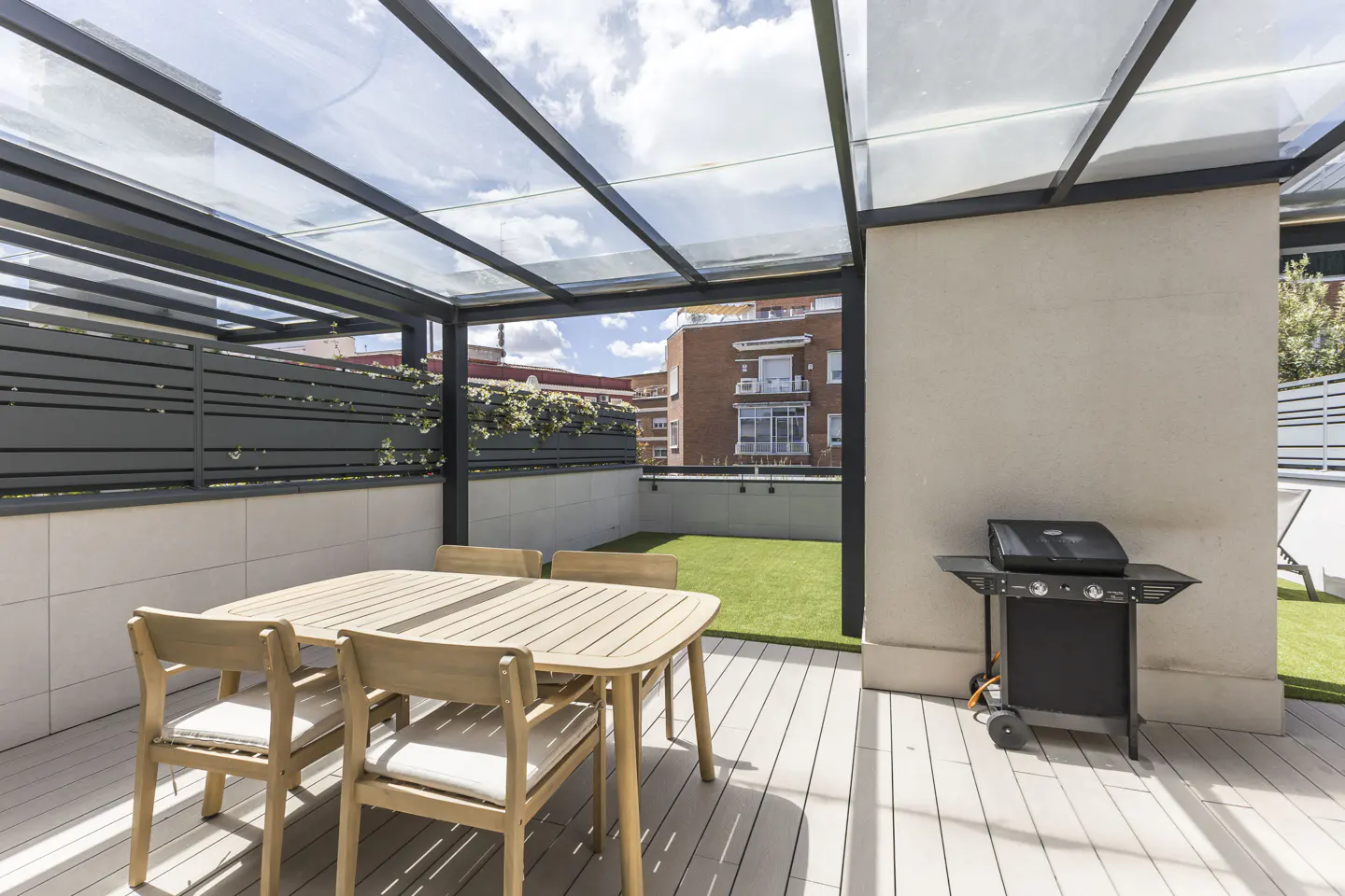 Outdoor patio with a glass roof, table, chairs, and grill. A building is visible in the background.