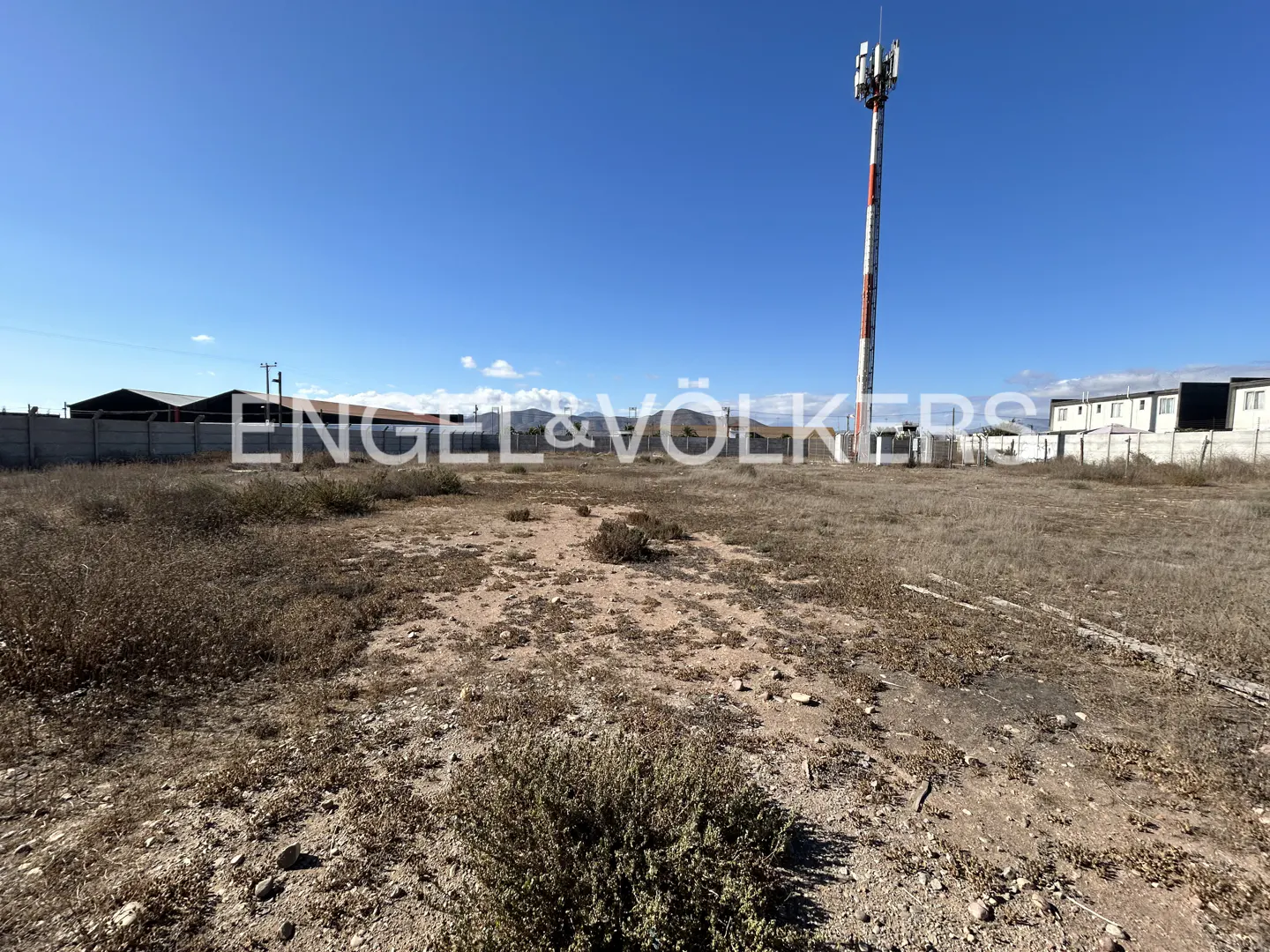 Vacant lot with dry brush under a blue sky. A red and white cell tower stands in the background. Engel & Völkers logo is superimposed.
