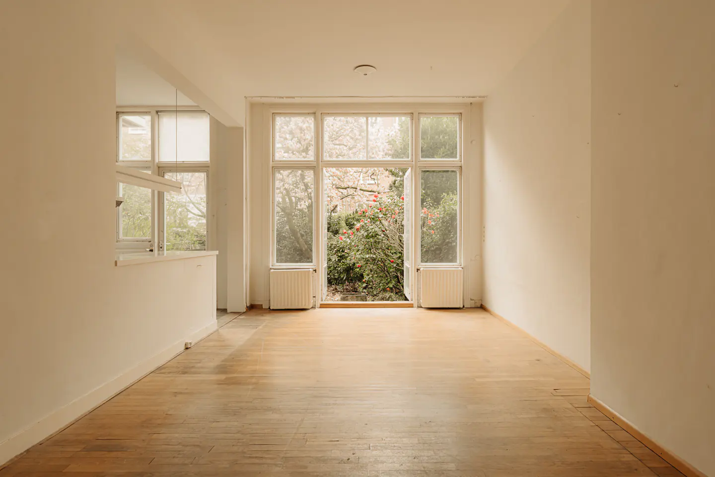Empty room with hardwood floors and white walls. Large open doors lead to a garden with green plants and red flowers.
