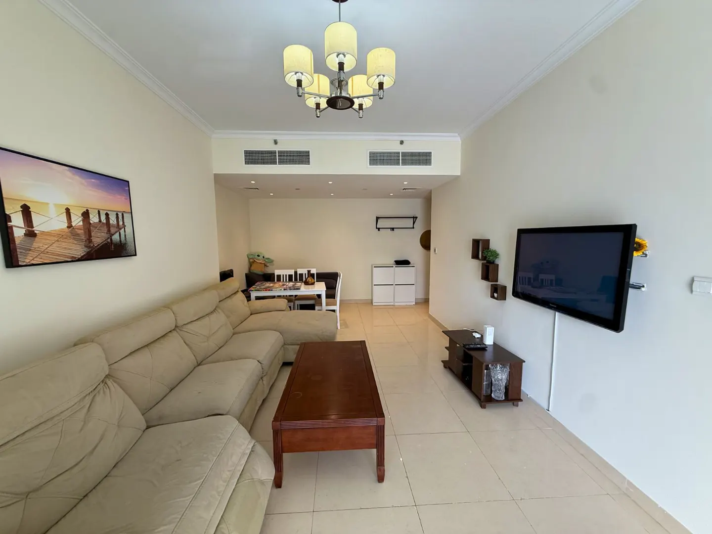 Living room with beige sectional sofa, wood coffee table, TV, and dining area. A beach scene art hangs on the wall.