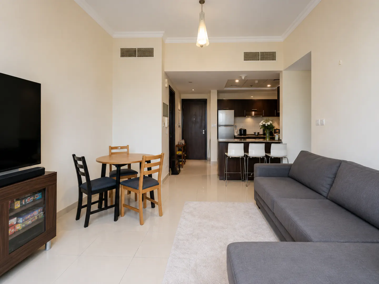 Open-concept apartment with a gray sofa, round wood table with chairs, and a kitchen with bar stools.