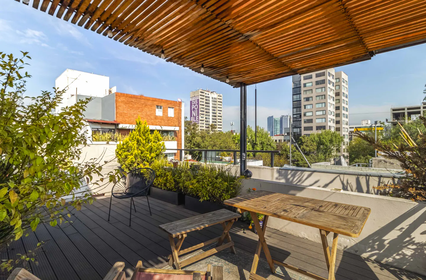 Rooftop patio with wooden table, bench, and chair. A wooden pergola provides shade. City buildings and greenery are in the background.