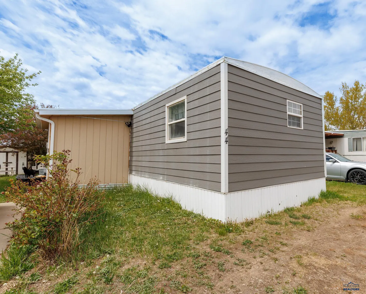 Exterior view of a gray and tan mobile home with white trim, set on a grassy lot under a blue sky with light clouds.