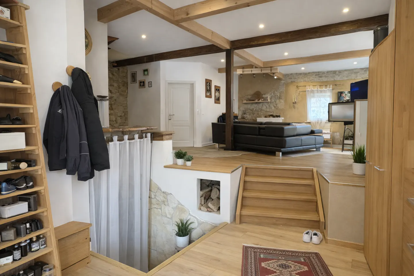 Open-plan living area with wooden beams, a sunken staircase, a black leather sofa, and a wooden shoe rack in the foreground.