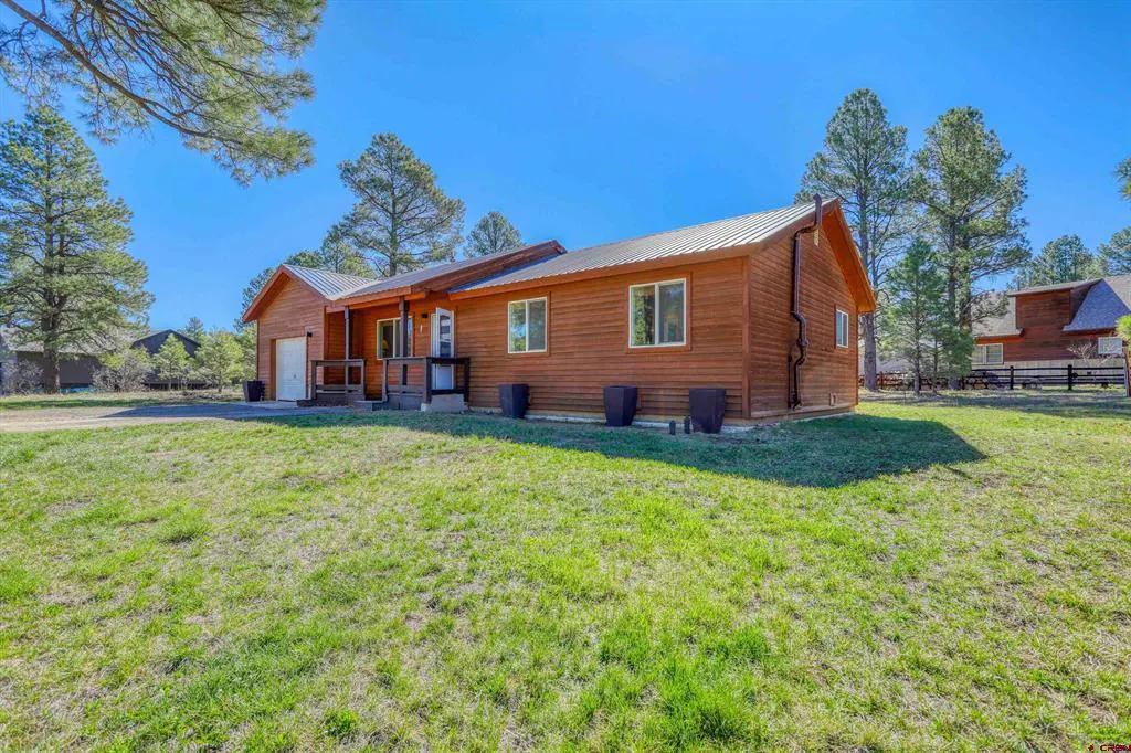 Exterior view of a one-story wood cabin with a metal roof, green lawn, and tall pine trees under a clear blue sky.