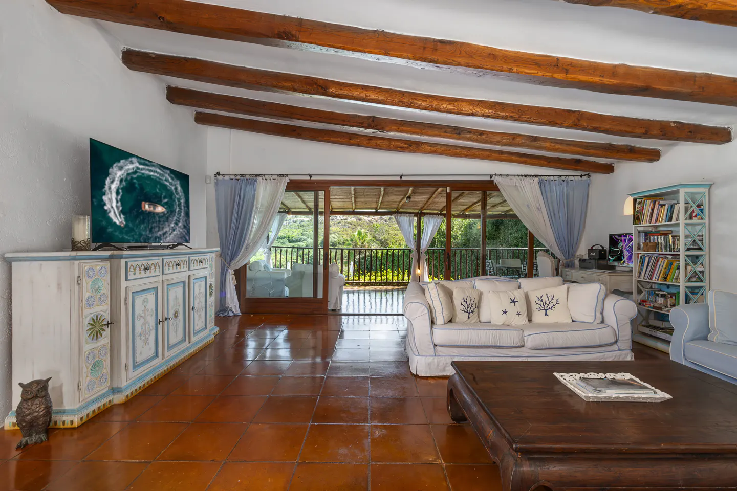 Bright living room with white walls, exposed wood beams, and terracotta tile floor. A white sofa with blue trim faces a dark wood coffee table. Sliding doors lead to a covered patio.