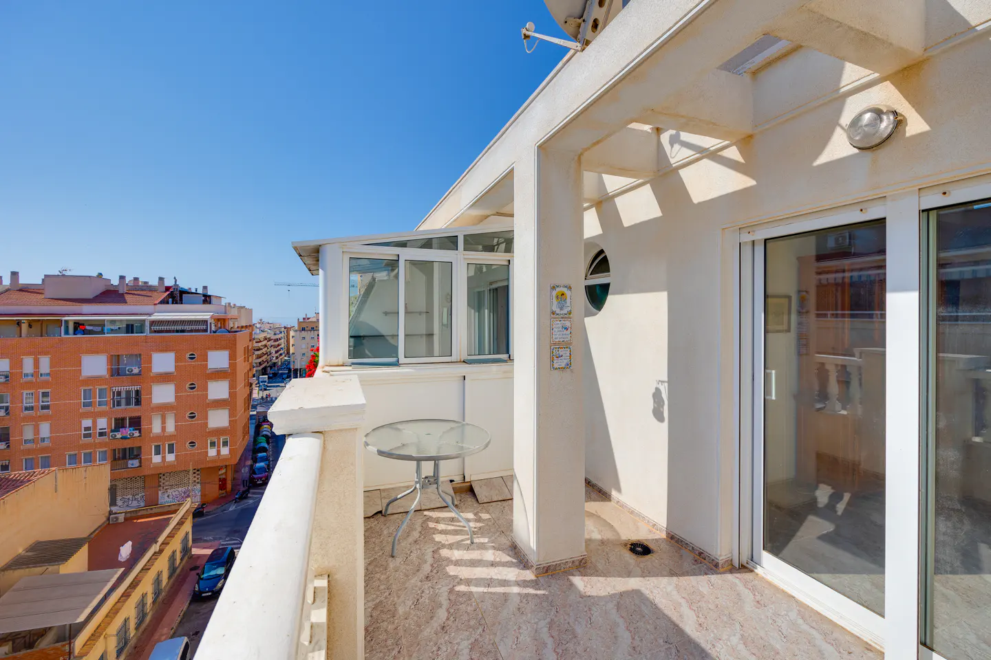 Sunny apartment balcony with a glass table, tiled floor, and a view of the surrounding city buildings.