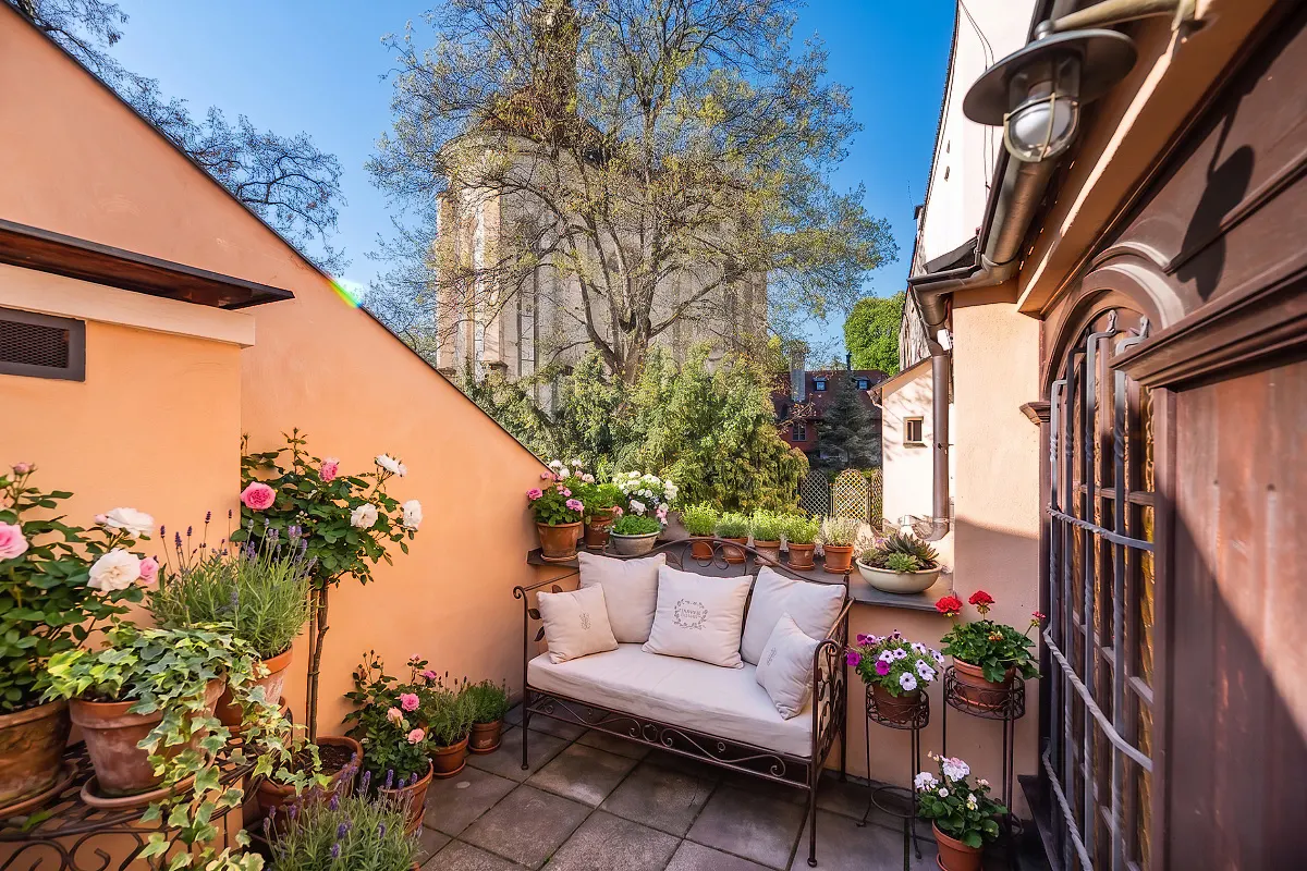 Cozy outdoor patio with a cream bench, potted flowers, and a view of a church tower against a blue sky.