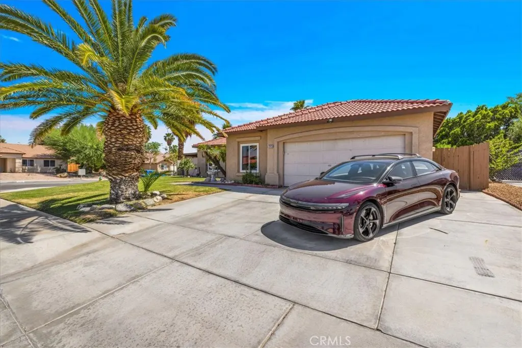 Tan single-story home with a red tile roof, a palm tree, and a maroon Lucid car in the driveway under a blue sky.
