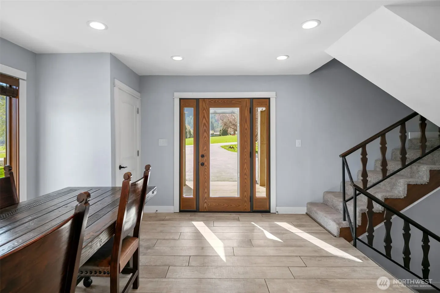 Entryway with wood-framed glass door, staircase, and dining table. Light gray walls and tile flooring. View of driveway and lawn through the door.