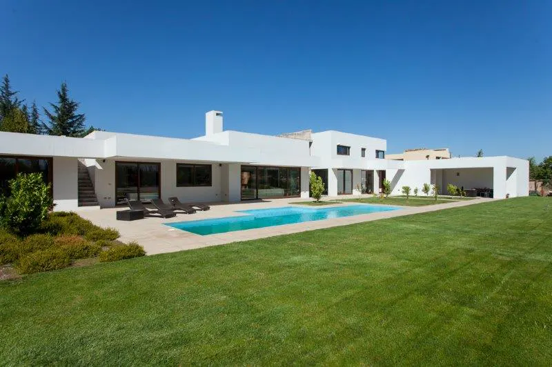Modern white house with a blue pool, green lawn, and lounge chairs under a clear blue sky.
