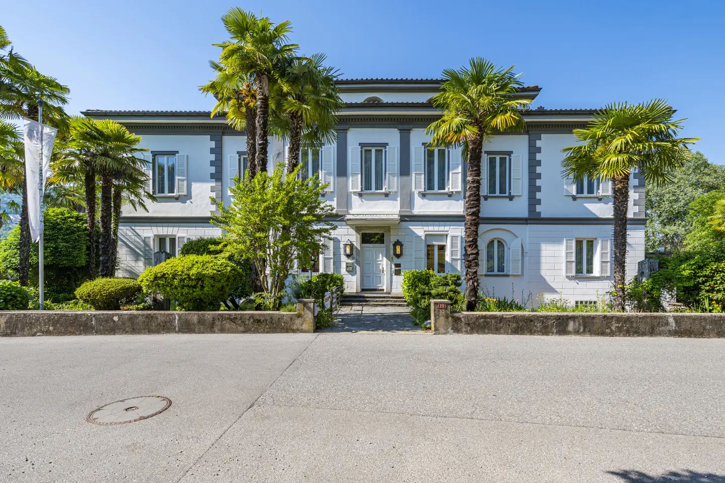 A white, two-story villa with grey trim, surrounded by palm trees and lush greenery under a clear blue sky.