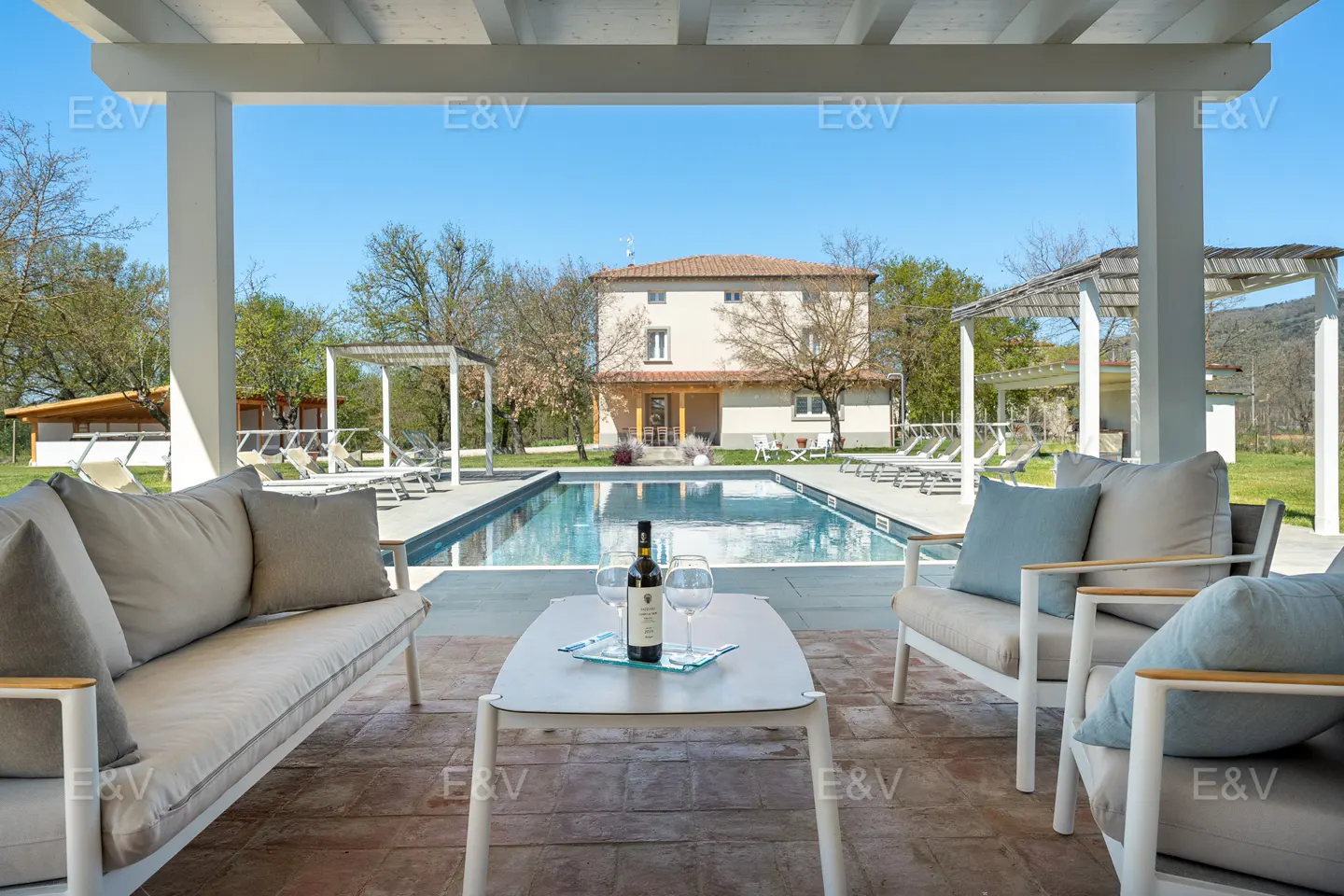 Outdoor patio with a pool view. Wine bottle and glasses on a table, with sofas and chairs. A house is in the background.