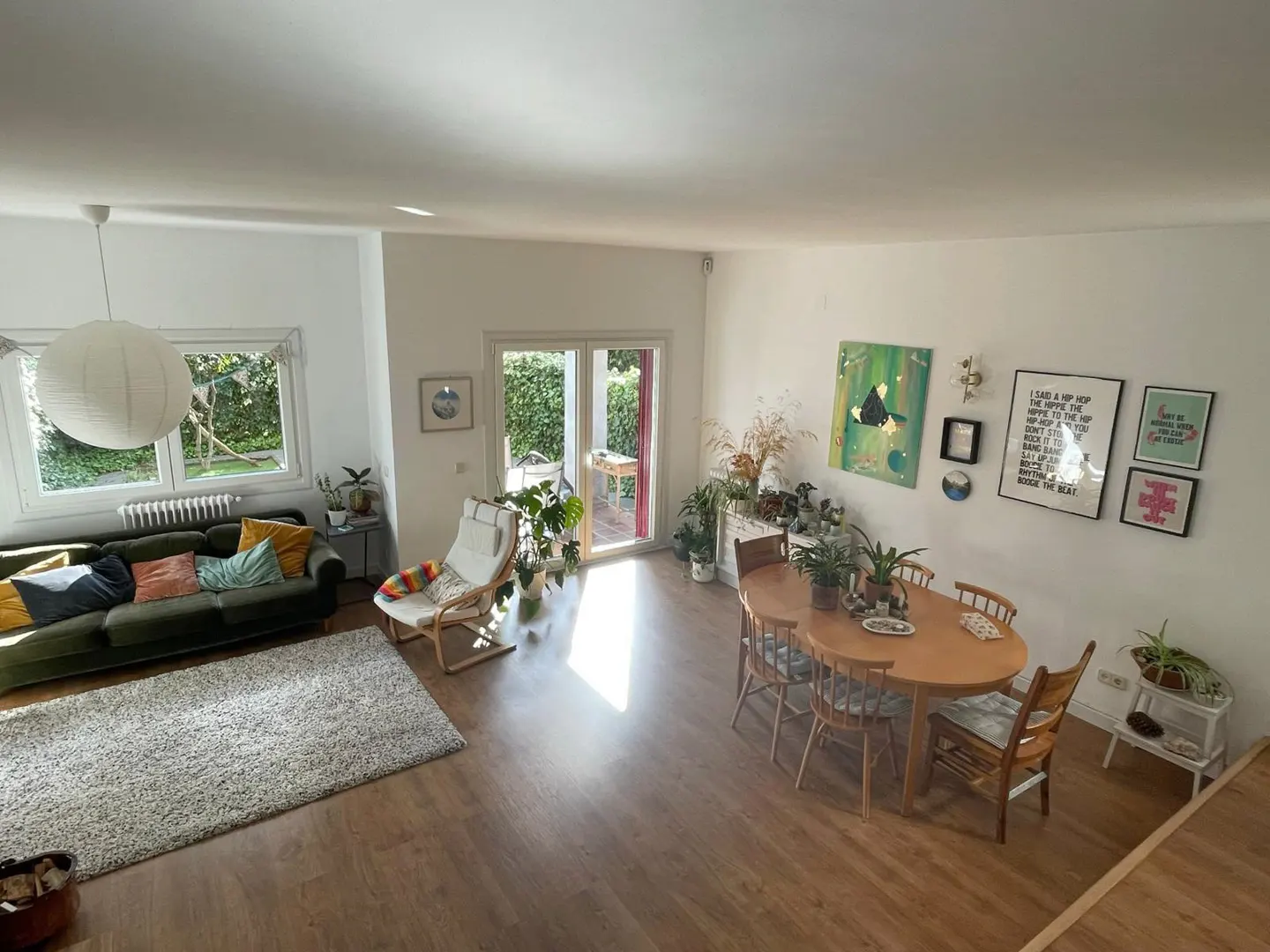 Bright, open-plan living space with wood floors, white walls, and large windows. A green sofa, dining table, plants, and art create a cozy atmosphere.