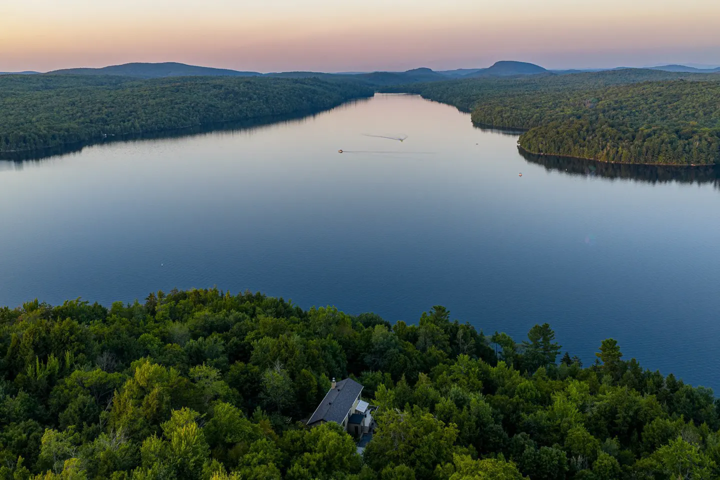 Aerial view of a lake surrounded by green forests, with a house nestled among the trees in the foreground.