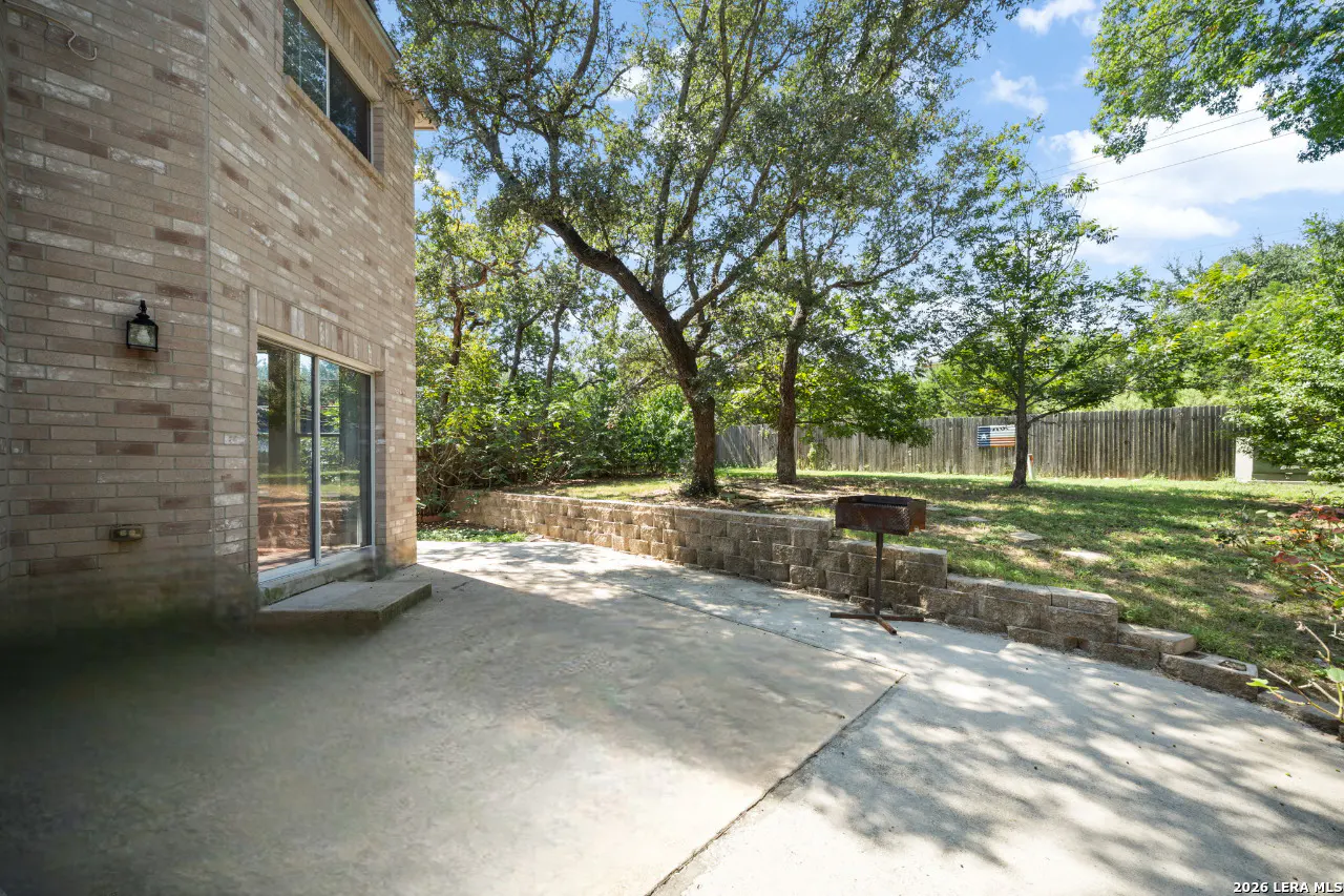 Backyard patio with brick house, sliding glass door, and stone retaining wall. Grill and trees in background.