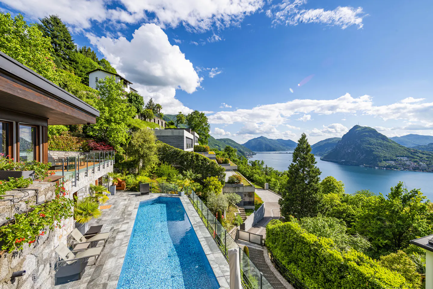 Luxury hillside home with a blue pool overlooking a lake and mountains under a blue sky with white clouds.
