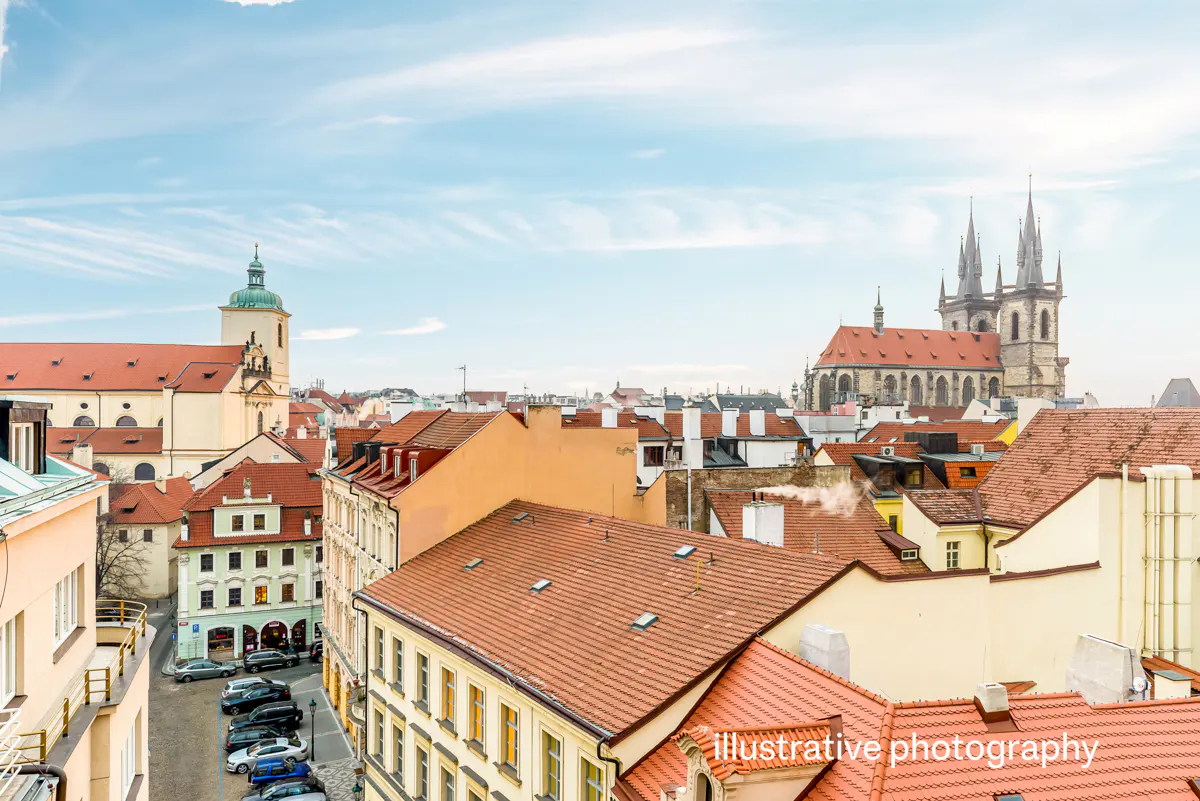 Cityscape view of Prague with red-tiled roofs, church towers, and a blue sky with light clouds.
