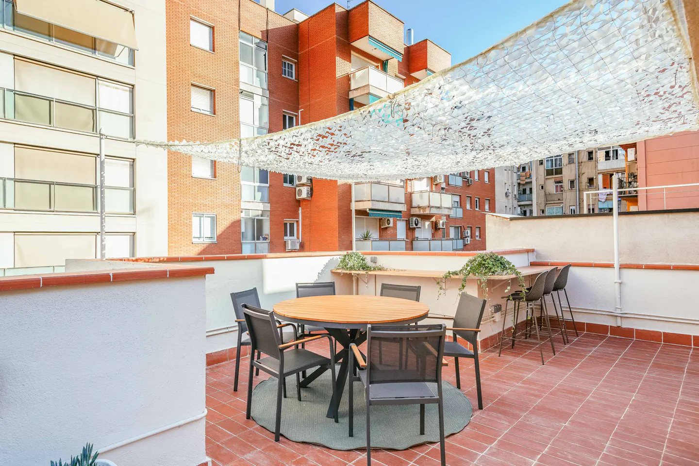 Rooftop patio with a round wooden table, six chairs, and a sunshade. Brick buildings are in the background.