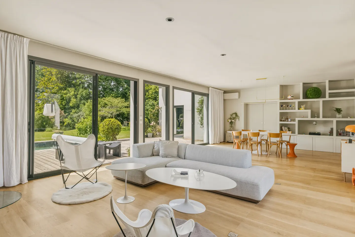 Bright, modern living room with a gray sectional sofa, white butterfly chairs, and large windows overlooking a green backyard with a pool.
