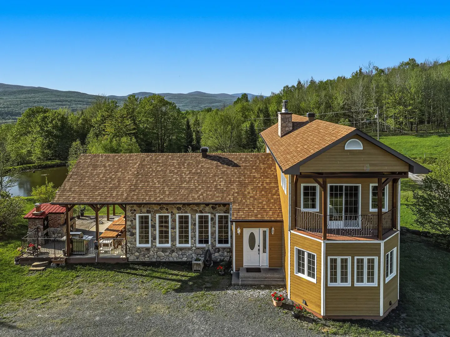 Two-story house with stone and yellow siding, brown roof, white trim, and a balcony, set against a backdrop of green trees and a blue sky.