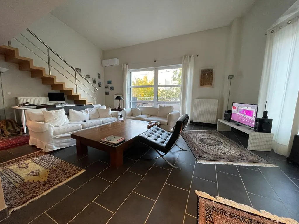 Bright living room with white sofas, a wooden coffee table, and a black leather chair on dark tile floors with rugs. A staircase is visible.