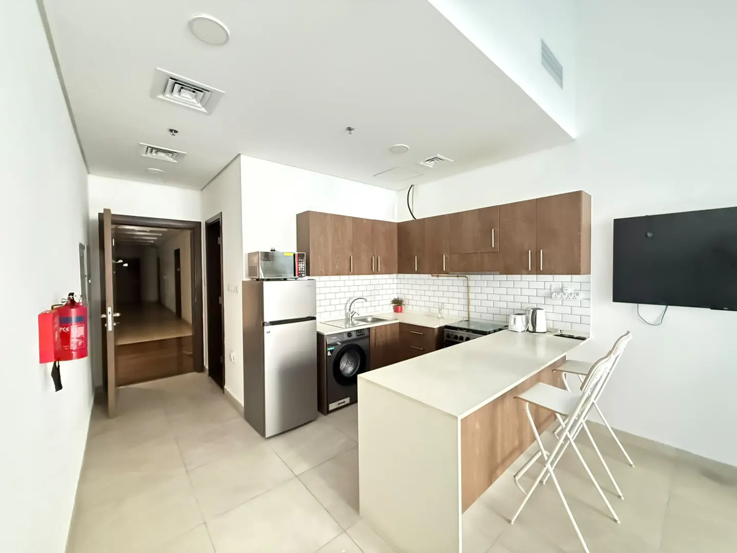 A bright, modern apartment kitchen with brown cabinets, white countertops, and stainless steel appliances. Two white chairs sit at the island. A hallway is visible in the background.