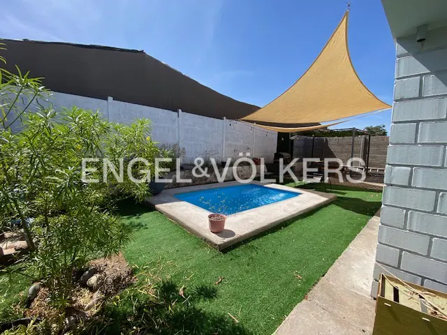 Backyard with a small blue pool, concrete surround, artificial turf, and a tan shade sail overhead. White fence and green foliage.