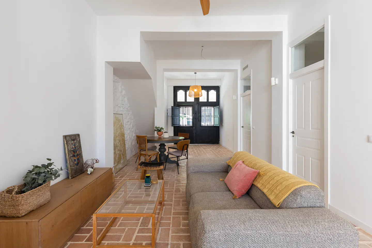 Bright living room with brick floors, white walls, and a gray sofa with pink pillow. A dining table and black doors are in the background.