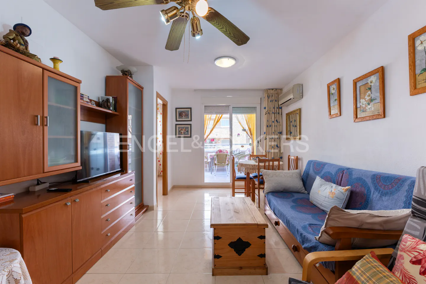 A bright living room with a blue sofa, wooden furniture, and a balcony view. The room has a ceiling fan and tiled floors.