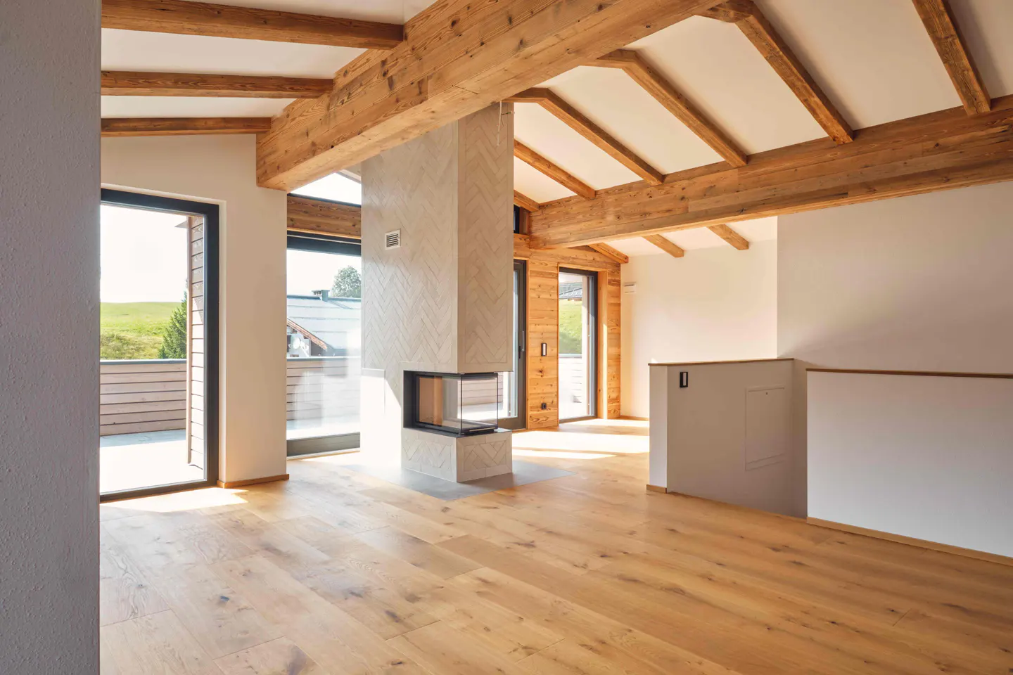 Bright, empty living room with wood floors, exposed beams, and a modern fireplace. Doors lead to a balcony with a green view.