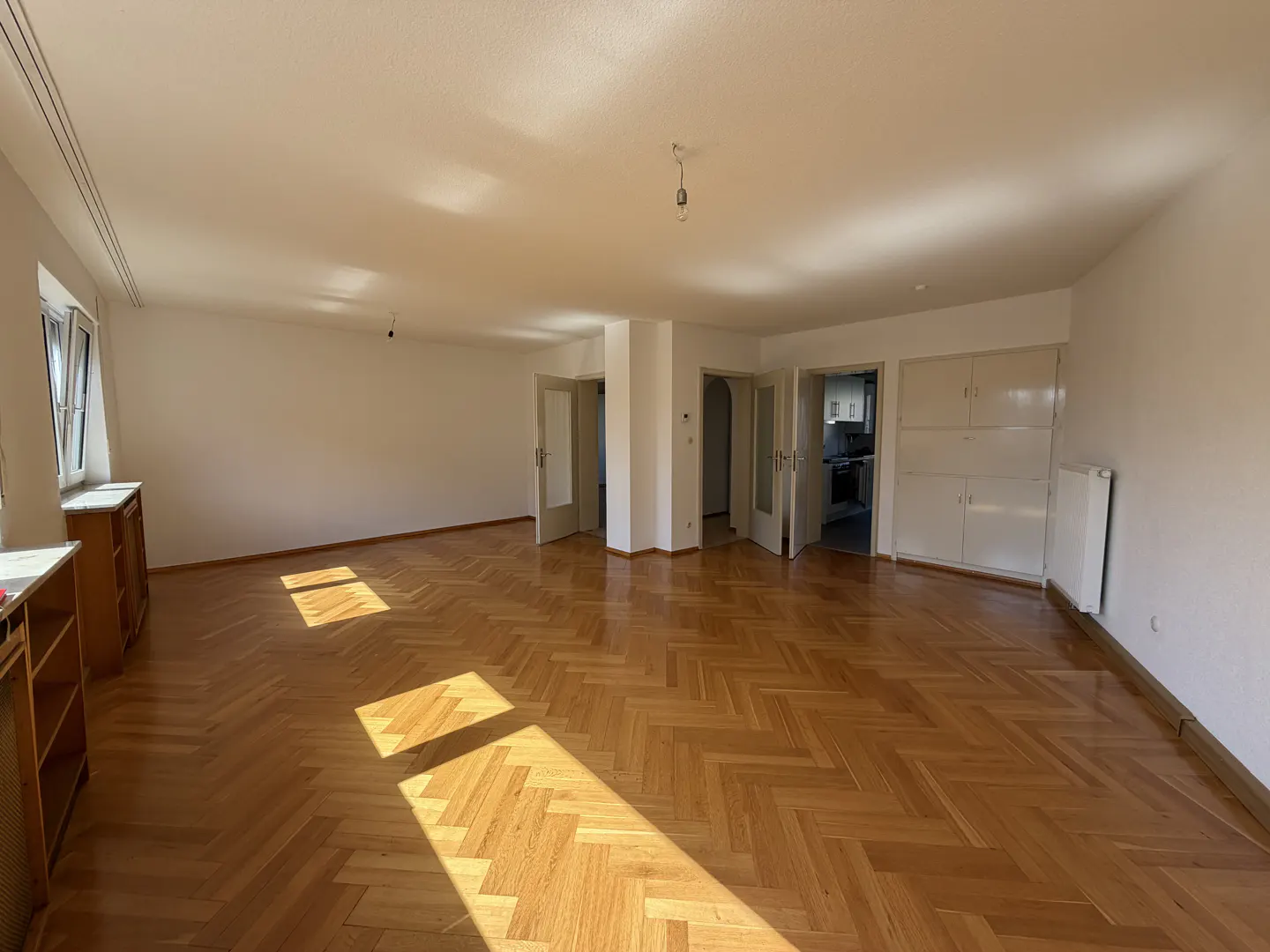 Spacious, empty room with light-colored walls, polished herringbone wood floors, and sunlight streaming through a window.