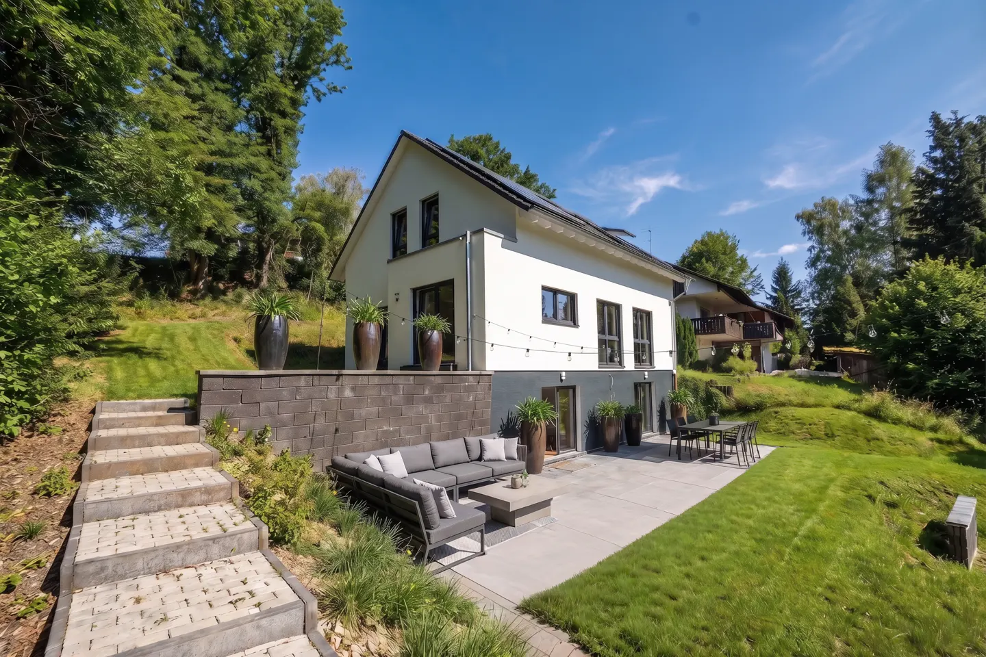 Two-story white house with a gray patio, outdoor seating, and a stone staircase leading up a grassy hill.
