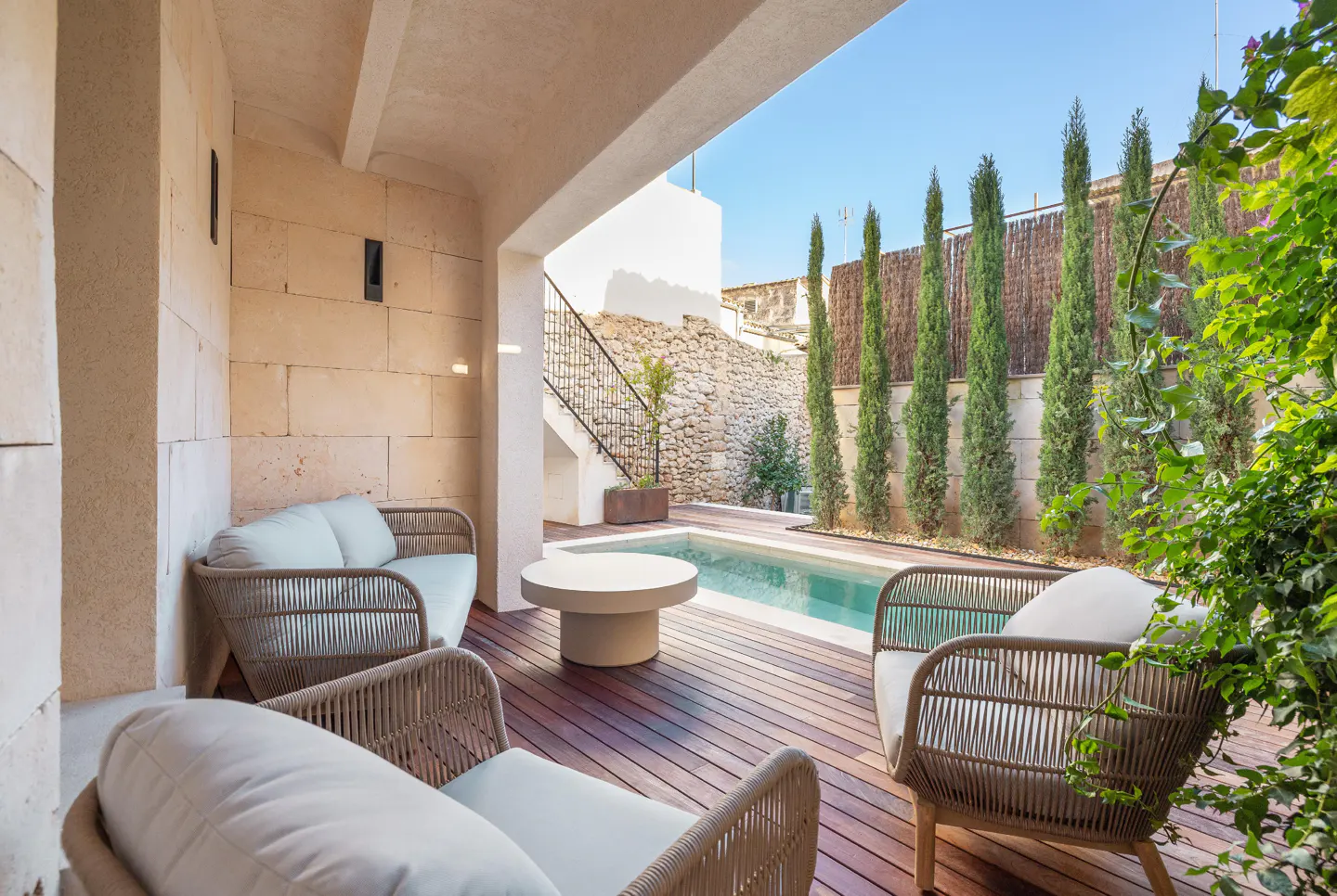 Outdoor patio with wicker furniture, a round table, and a pool surrounded by tall trees and a stone wall.