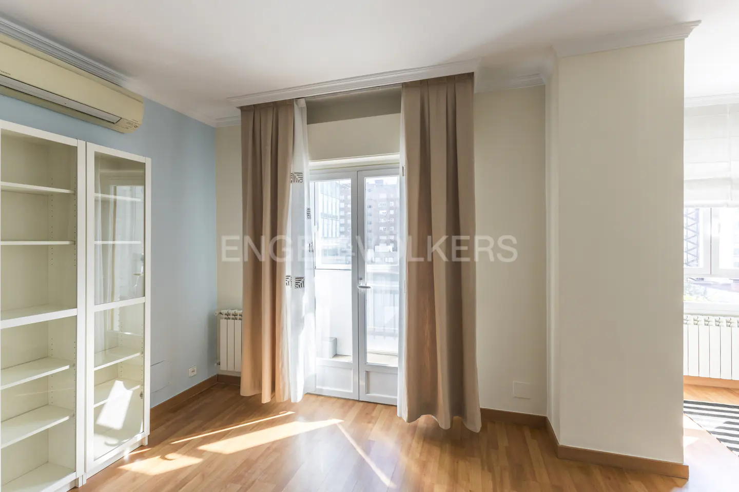 Bright living room with wood floors, white bookcase, and balcony doors with beige curtains.