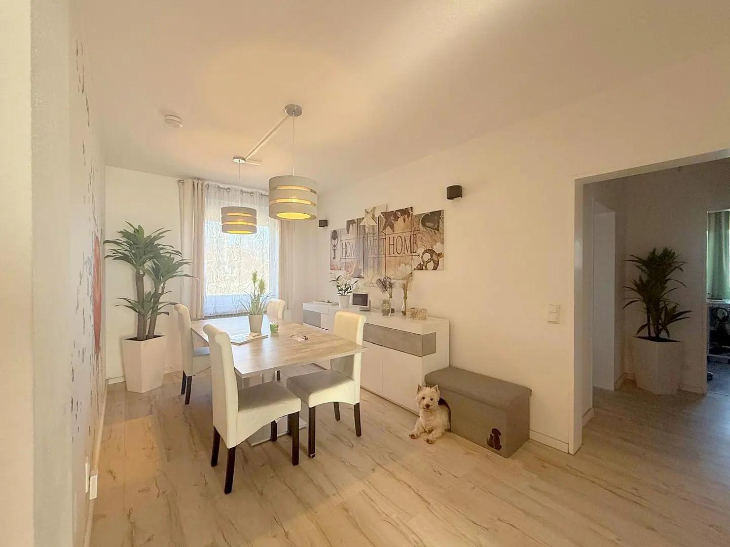 A bright dining room with a wooden table, white chairs, and a sideboard. A small white dog sits near a gray storage bench with a dog silhouette.