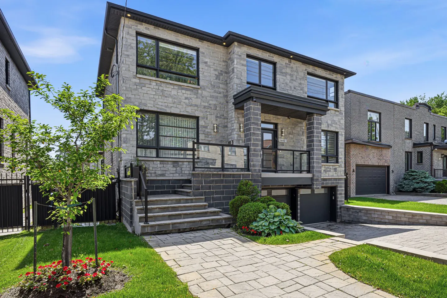 Modern two-story stone house with a paved walkway, front porch, and attached garage under a clear blue sky.